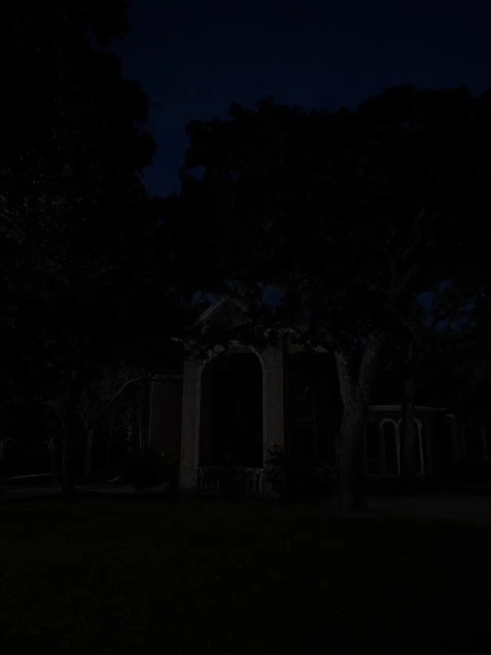 Brick two-story home with arched entry under oak trees before lighting installation
